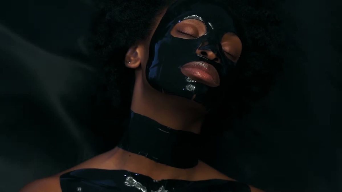 Model in a black 111SKIN face and neck mask lying on black satin
