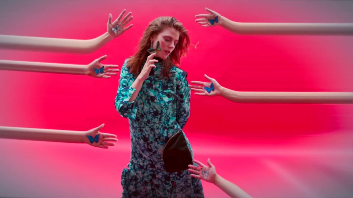 Rose Leslie in a printed dress surrounded by reaching hands on a pink set