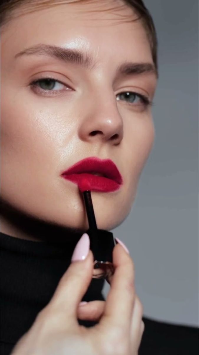 Model applying red lipstick in a black turtleneck against a grey backdrop
