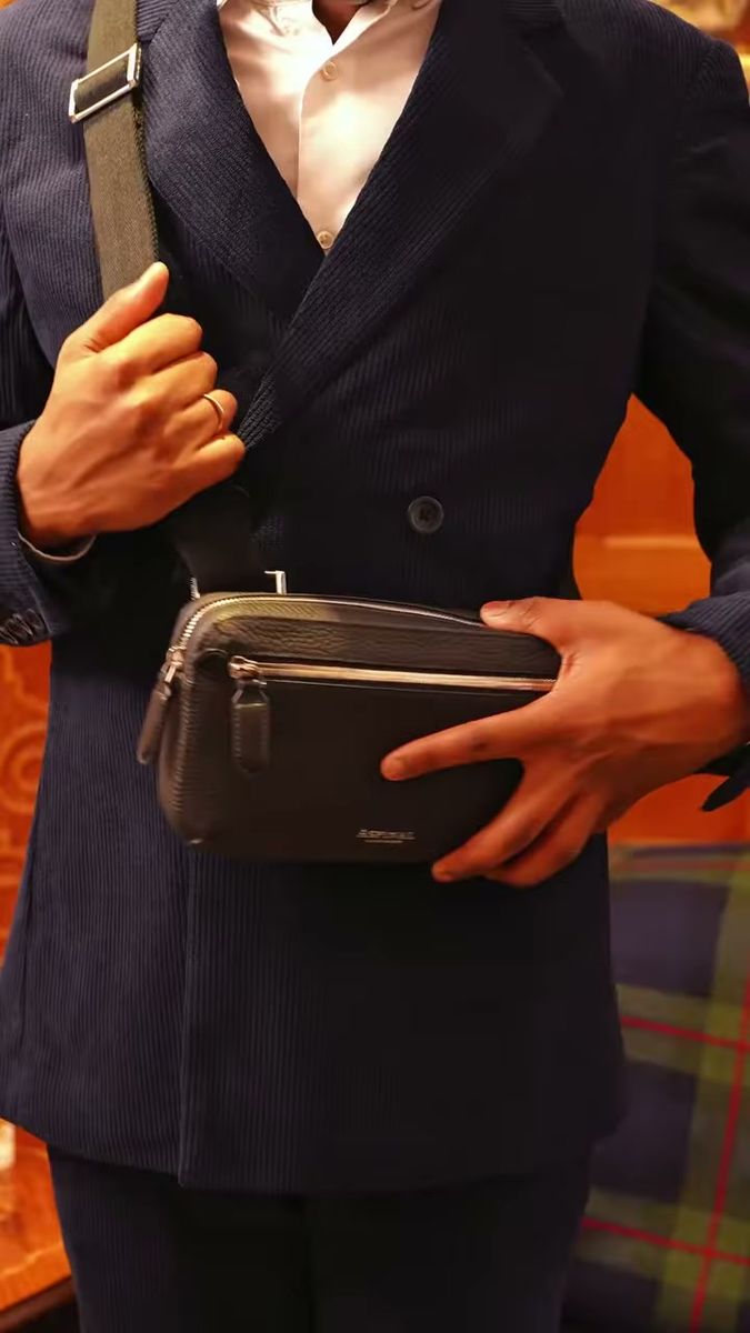 Hands on a black leather crossbody pouch over a navy double-breasted coat