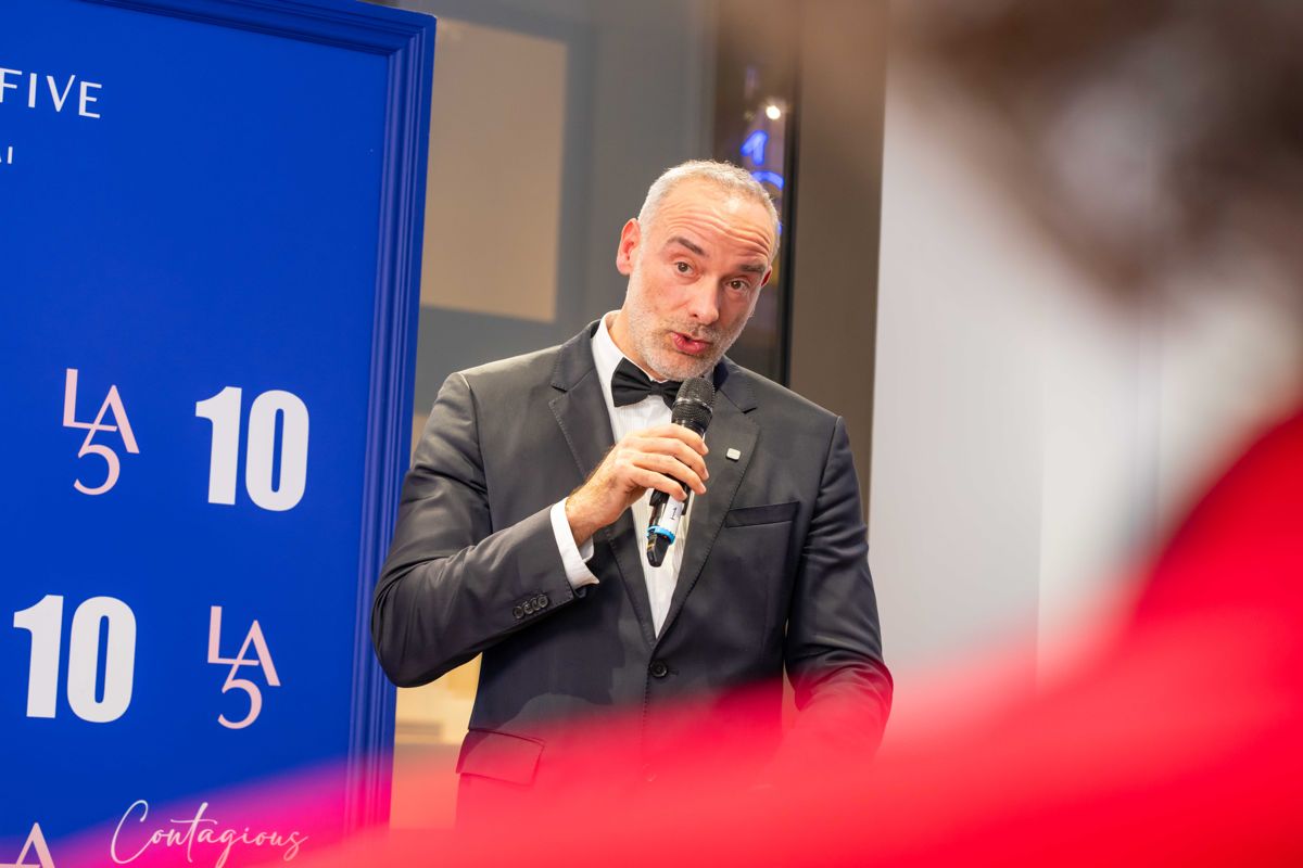 Speaker in a dinner jacket talking into a handheld microphone beside the L'Atelier Five backdrop at Opera Gallery London