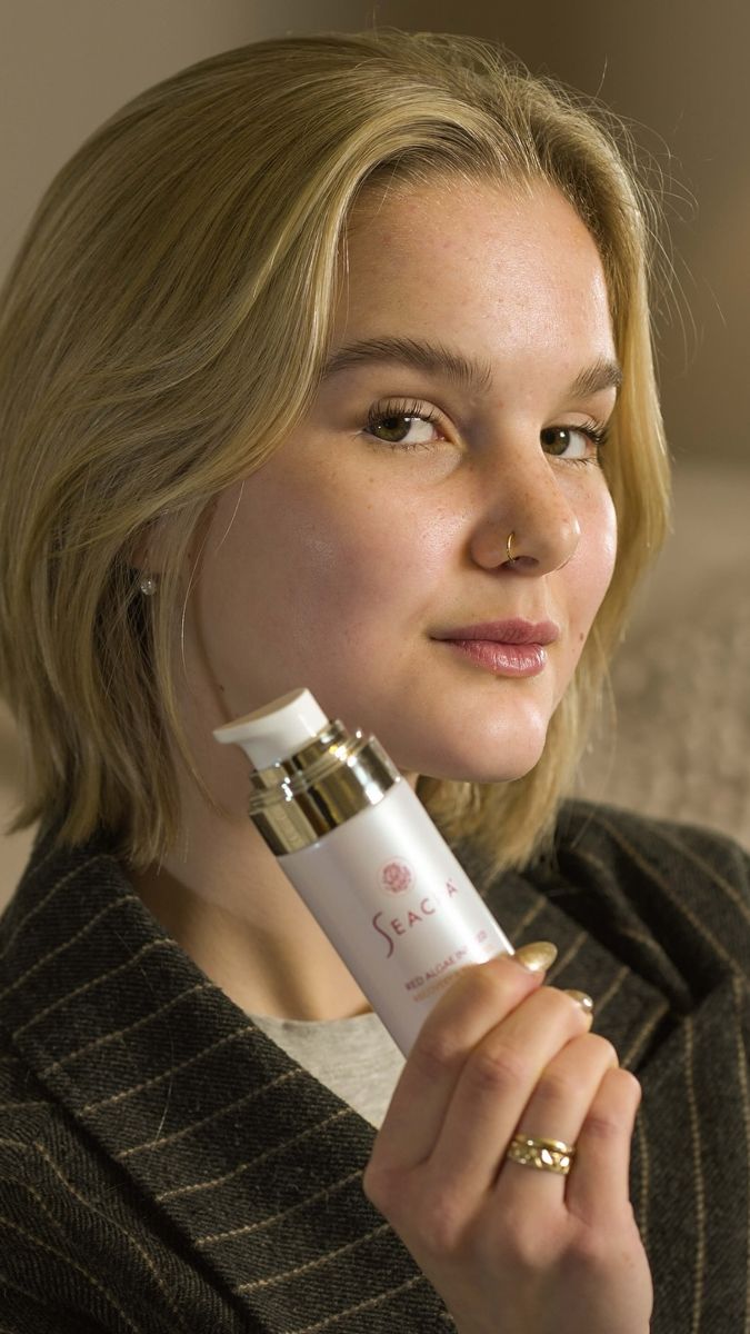 Blonde woman in a tweed jacket holding a Seacra bottle to her cheek, soft studio light