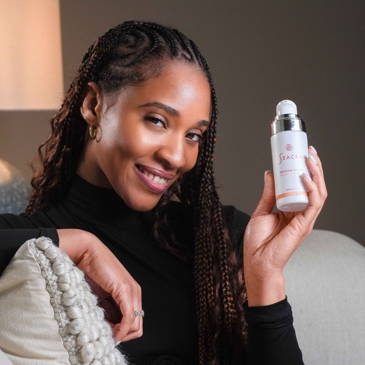 Woman with braided hair smiling on a sofa, holding a Seacra skincare bottle