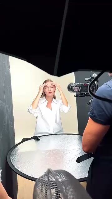 Video camera with a top monitor filming a model in a white studio, with a silver reflector table in front