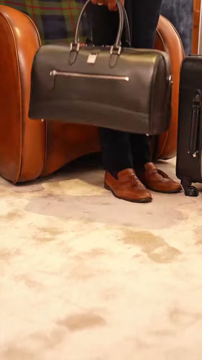 Aspinal of London black leather holdall set down by a tan leather armchair in a wood-panelled room