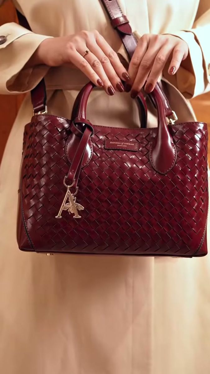 Hands with burgundy nails holding an Aspinal of London woven tote against a camel trench coat