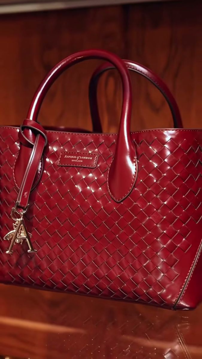 Aspinal of London burgundy woven leather tote with a gold A charm on a polished wooden shelf