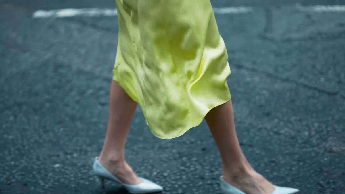 Silver pointed heels and a yellow satin dress mid-stride on asphalt