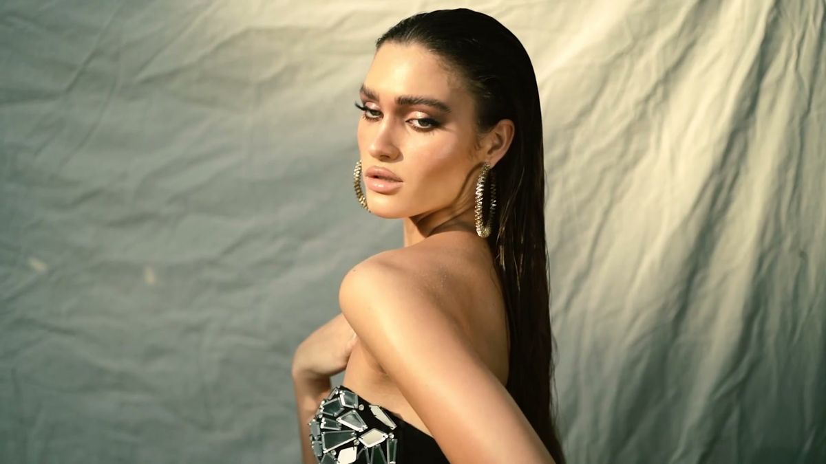 Model in a mirrored top looking over her shoulder against a cream fabric backdrop in golden light